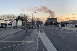 Eine Stadtstraße mit Absperrbaken, einem Bus, Laternenpfählen, Bäumen, Gebäuden, Kränen und Rauch, der aus einem entfernten Gebäude aufsteigt, vor einem Himmel als Hintergrund.