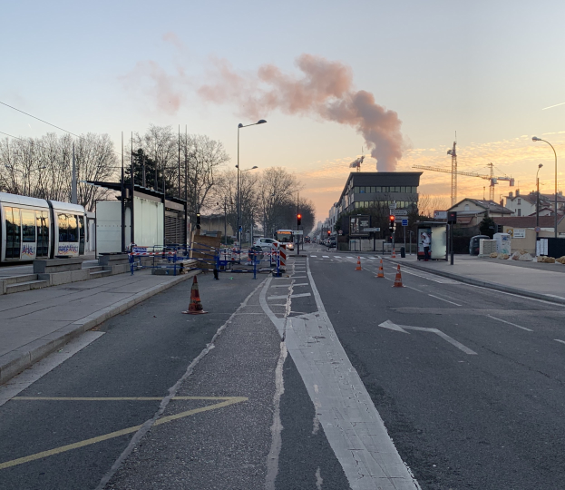 Eine Stadtstraße mit Absperrbaken, einem Bus, Laternenpfählen, Bäumen, Gebäuden, Kränen und Rauch, der aus einem entfernten Gebäude aufsteigt, vor einem Himmel als Hintergrund.