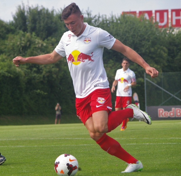 Ein professioneller Fussballspieler in rot-weißer Uniform tritt einen Fussball auf einem grünen Feld mit Bäumen und einem klaren blauen Himmel im Hintergrund, wobei ein "RB Leipzig"-Schild sichtbar ist.