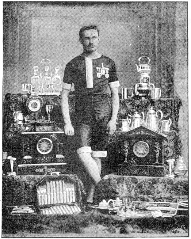 Schwarzes und weißes Foto eines Mannes in einem Fußballtrikot, der vor einem Tisch mit Pokalen, Trophäen und anderen Gegenständen steht, mit Text unten: "1909-1910 Weltmeisterschaftssieger."