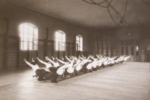 Gruppe von Menschen, die Pilates-Übungen in einem Fitnessstudio mit Fenstern und einer Uhr an der Wand durchführt.
