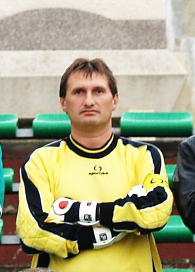 Ein Torwart in einem gelben Trikot steht neben einem anderen Mann in einer schwarzen Jacke, mit grünen Stühlen und einer Wand im Hintergrund.