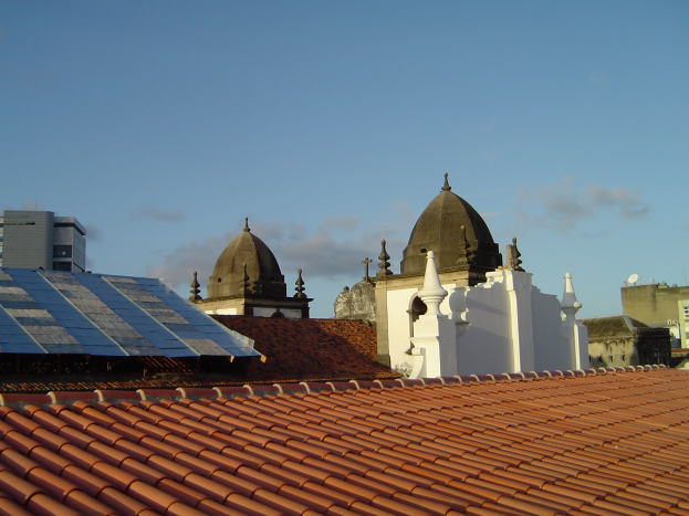 Eine Stadtansicht mit einigen Gebäuden im Vordergrund, von denen eines Solarpaneele auf dem Dach hat, unter einem blauen Himmel.