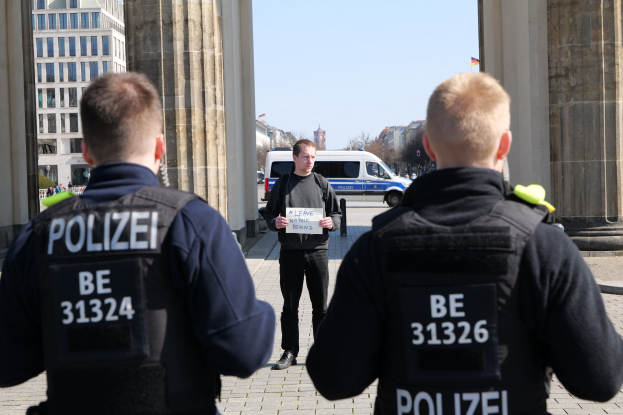 Drei Polizeibeamte stehen vor einem Gebäude, ein Mann hält ein Papier zwischen ihnen, flankiert von Säulen und Fahrzeugen im Hintergrund.