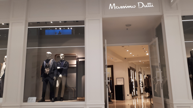 Vitrine des Massimo Dutti in Mailand mit Schaufensterpuppen in moderner Kleidung, beleuchteter Decke, Informationsschildern und sichtbarem Boden.