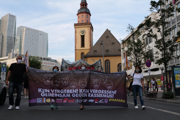 Eine Gruppe von Menschen in Masken, die eine Banner mit Text und Bildern tragen, geht eine Straße entlang, mit einem geparkten Auto auf der linken Seite, Gebäuden, Bäumen, Schildern, Polen, einem Uhrenturm und einem klaren blauen Himmel im Hintergrund.
