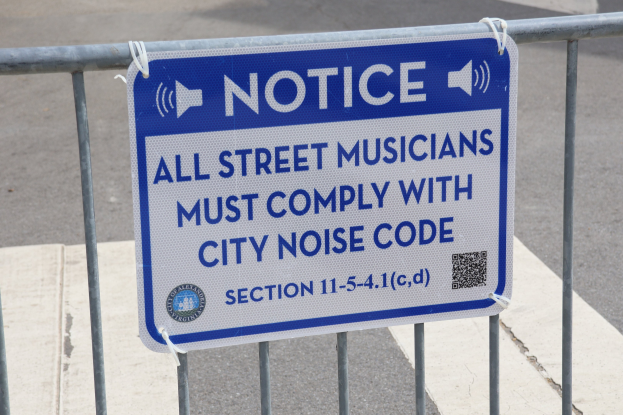 Anschlagtafel an einem Zaun mit einem Schild, auf dem "Anschlag: Alle Straßenmusikanten müssen die Stadtlärmvorschrift einhalten" steht, und eine Straße im Hintergrund.