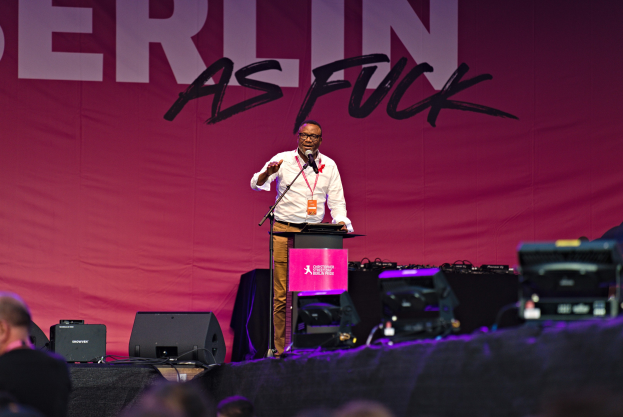 Mann in weißem Hemd und braunen Hosen, der in ein Mikrofon spricht, vor einem pinken 'Berlin as Fuck'-Plakat.