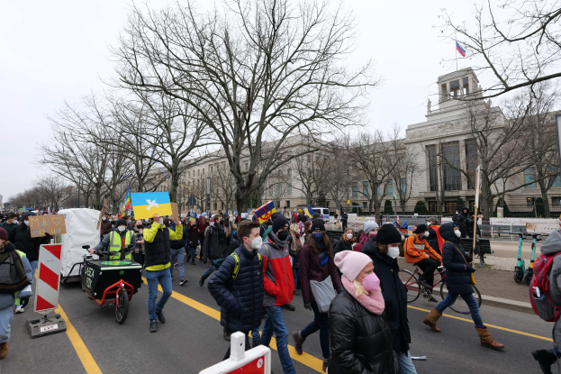 Ein große Demonstrationszug mit Menschen, die eine Straße in Washington, D.C., entlanggehen, einige halten Schilder und andere fahren Fahrräder, mit Bäumen und einem klaren blauen Himmel im Hintergrund.