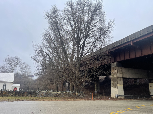 Eine Brücke unter Reparatur überspannt eine Straße, mit einem zentralen Baum umgeben von Steinen und Gras, einem nahen Haus, Schild, Straßenpfahl und Gruppe von Bäumen, vor einem bewölkten Himmel.