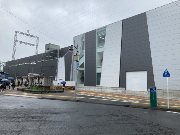 Großes mehrstöckiges Gebäude mit zahlreichen Fenstern, Straßeninfrastruktur einschließlich Pfählen und Kabeln, Fahrzeuge auf der Straße, Fußgänger mit Schirmen auf dem Gehweg, Bäume und ein bewölkter Himmel, identifiziert als das Tokyo Metropolitan Government Building.
