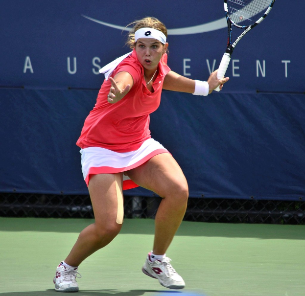 Eine Frau in einem pinken Shirt und weißen Schuhen, die ein Headband trägt, spielt Badminton auf einem Court und hält einen Schläger, wobei ein Banner im Hintergrund zu sehen ist.