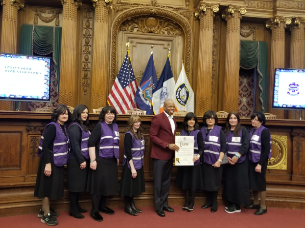 Gruppe von Menschen vor Flaggen mit einem Mann in der Mitte, der ein Zertifikat hält, flankiert von zwei Bildschirmen, vor einer hölzernen Wand mit kunstvollen Schnitzereien und Vorhängen, was auf eine Stadtratssitzung hinweist.