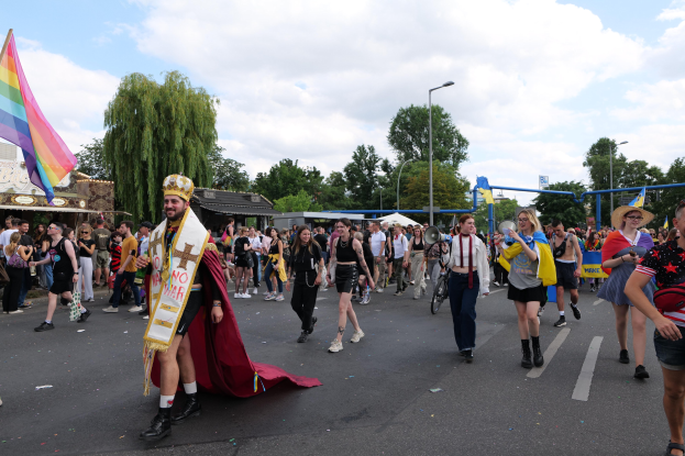 Eine Gruppe von Menschen marschiert bei der Gay Pride Parade 2018 mit einer Regenbogenflagge und Musikinstrumenten, mit Laternenmasten, Bäumen und Hütten im Hintergrund.