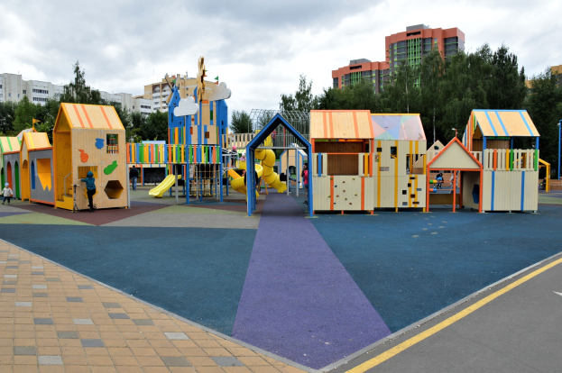 Kinderspielplatz in einer städtischen Umgebung mit Gebäuden, Bäumen und einem bewölkten Himmel, mit einigen Menschen und umgeben von einer Straße.