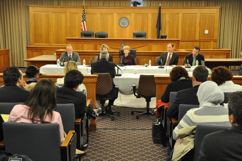 Ein Versammlungsraum mit Menschen auf Stühlen, zwei ausländische Flaggen, ein oranger Schreibtisch mit Wasserflaschen und einem Mikrofon.