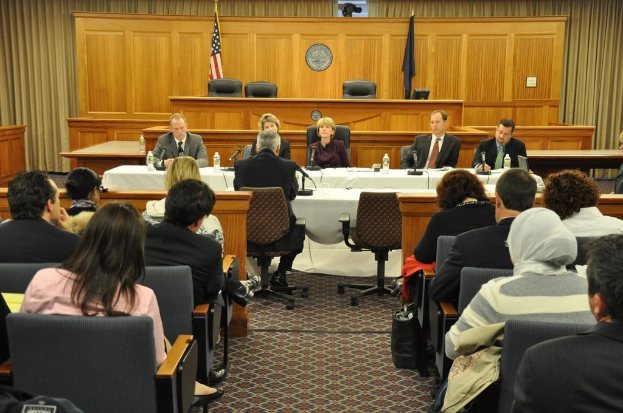 Ein Versammlungsraum mit Menschen auf Stühlen, zwei ausländische Flaggen, ein oranger Schreibtisch mit Wasserflaschen und einem Mikrofon.