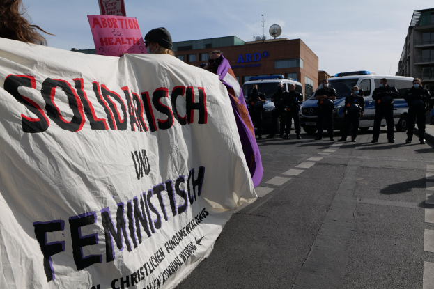 Eine Gruppe von Menschen marschiert auf einer Straße, hält ein Banner mit der Aufschrift "Solidarität und Feminismus" in der Hand, mit geparkten Fahrzeugen am Straßenrand und Gebäuden im Hintergrund unter einem klaren blauen Himmel.