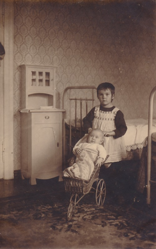 Ein altes Foto von zwei Kindern, die in einem Raum mit einem Baby im Kinderwagen, einem Bett im Hintergrund, einem Schrank zur Seite und einem Teppichboden stehen.