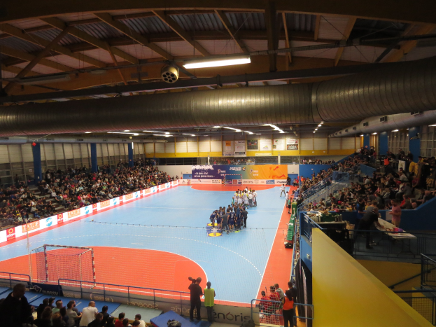 Große Halle mit Zuschauern bei einem Handballspiel, mit Sitzplätzen und Stehplätzen, einem Netz auf der linken Seite, Texttafeln im Hintergrund und Beleuchtung und Rohren an der Decke.