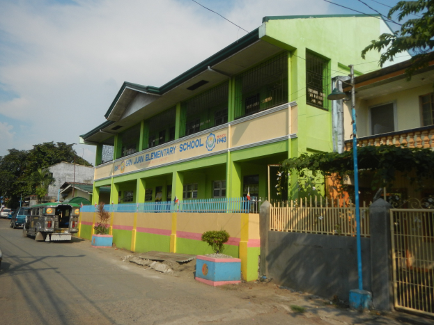 Schulgebäude mit grüner und gelber Fassade, Straße mit Fahrzeugen, Topfpflanzen, Zaun, Tor, Straßenlaterne, Bäume, Kabel und bewölktem Himmel.
