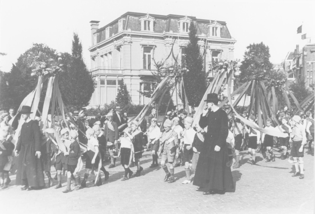 Gruppe von Menschen, die eine Straße entlanggehen und Fahnen vor einem Gebäude schwenken, tragen Hüte, mit Maibaum, Bäumen, Gebäuden und Himmel im Schwarzweiß.