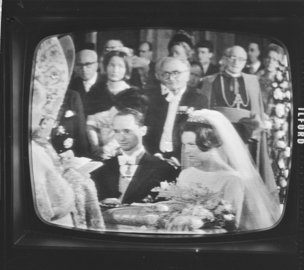 Schwarzes und weißes Foto einer Braut in einem weißen Kleid und Schleier, die einen Strauß hält, neben einem Bräutigam in Anzug und Krawatte steht, auf einem Fernsehbildschirm umgeben von einer Gruppe von Menschen.