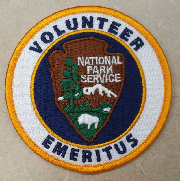 Runder Patch mit "Volunteer National Park Service Emeritus" in fetter schwarzer Schrift auf einem weißen Hintergrund, umrandet von einem blauen Rahmen und einem gelben Kreis.
