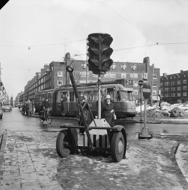 Schwarzes und weißes Foto einer Ampel an einer Kreuzung mit Fahrzeugen, einem Radfahrer, Passanten, Gebäuden, Strommästen und Drähten im Hintergrund.