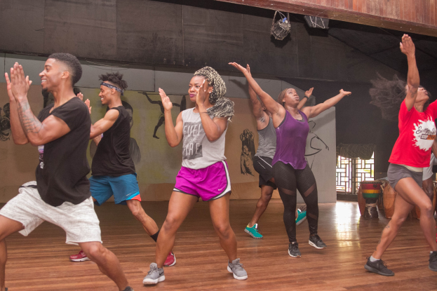 Gruppe von Menschen, die in einem Studio mit einer bemalten Wand, einem Fenster und Deckenlampen tanzen, wahrscheinlich während eines Zumba-Kurses.