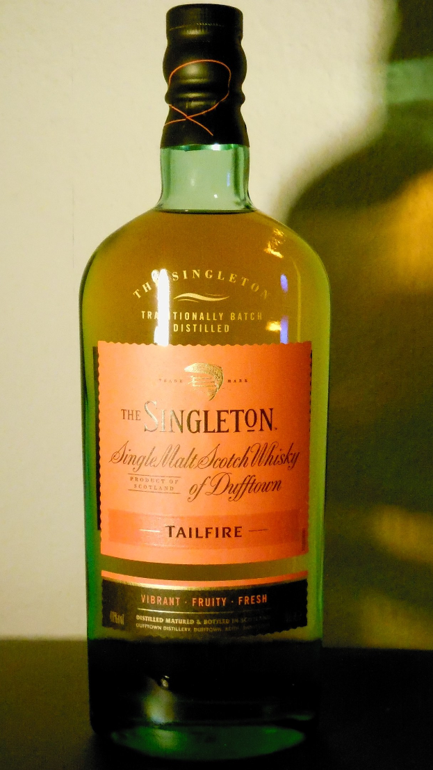 Eine tiefe Bernsteinflasche Singleton of Dufftown Tailfire Single Malt Scotch Whisky mit einem schwarzen Etikett, das ein Löwen- und Schlosswappen zeigt, auf einem Tisch.