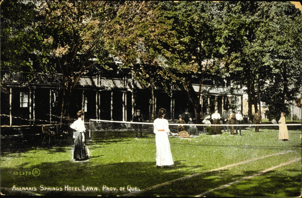 Schwarz-weiß-Foto einer Gruppe von Menschen, die Tennis auf dem Rasen vor dem Abenakis Springs Hotel Lawn in Provo, Quebec spielen, mit einem Netz, Stühlen, B├Ąumen und einem Geb├Ąude im Hintergrund.