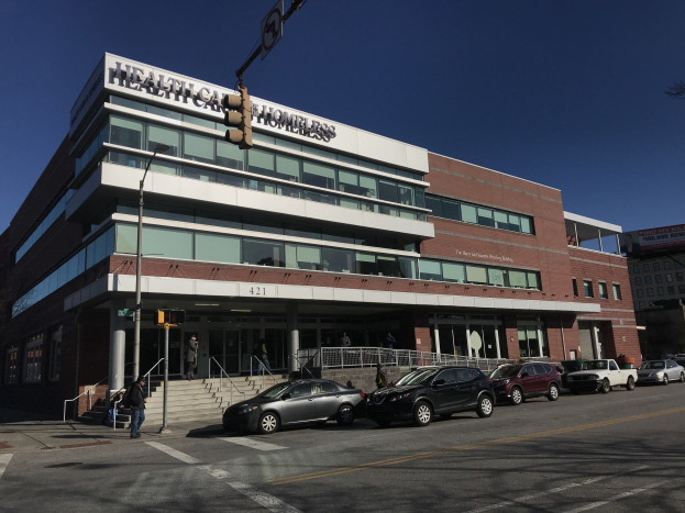 Großes Gebäude mit der Aufschrift 'New York City Health Care Homeless Center', vor dem Autos geparkt sind, umgeben von Straßeninfrastruktur und Passanten unter einem klaren blauen Himmel.