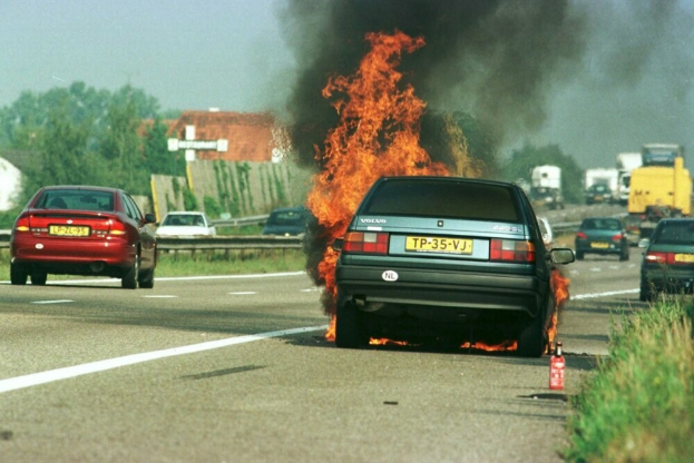 Ein Auto, das in Flammen steht, umgeben von anderen Fahrzeugen, mit Bäumen, Gebäuden und einem klaren blauen Himmel im Hintergrund, Gras auf der rechten Seite und ein Feuerlöscher in der Nähe.