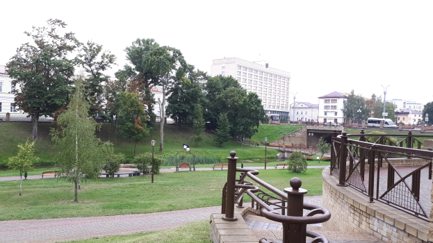 Ein Park mit einer Brücke über einen Fluss, umgeben von Gebäuden, Bäumen, Straßenlaternen, Straßenlaternen, Kraftfahrzeugen, Geländern, Bänken, Pflanzen, Gras und einem klaren blauen Himmel.