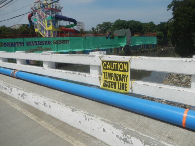 Eine Brücke mit einem "Vorsicht temporäre Wasserleitung"-Schild, Geländern, Bäumen, Gebäuden, einer Wasserrutsche und einem bewölkten Himmel im Hintergrund.