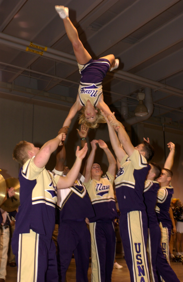 Eine Gruppe von Cheerleadern führt einen Stunt in einer Turnhalle durch, wobei eine Frau in einem Handstand in der Mitte steht, andere um sie herum stehen, einige halten Instrumente auf der linken Seite und eine Wand mit Rohren und Deckenleuchten im Hintergrund zu sehen ist.