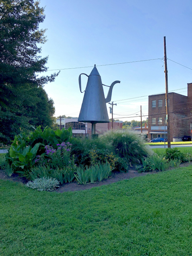 Ein Metallgießer auf einem grünen Feld mit Pflanzen, Bäumen, Strommasten, einem Schild, Fahrzeugen, Gebäuden und einem bewölkten Himmel im Hintergrund.
