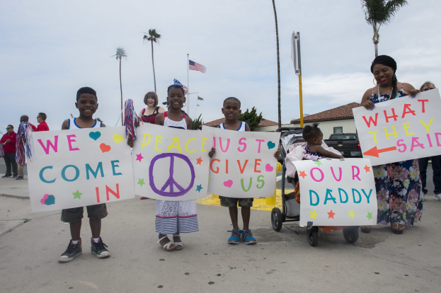 Eine Gruppe von Kindern hält Schilder in der Hand und steht vor einem Gebäude, während eine Frau einen Kinderwagen mit einem Baby schiebt.