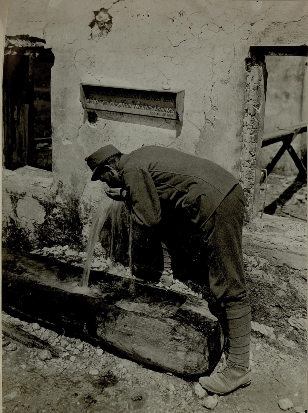 Ein altes Schwarz-Weiß-Foto eines Mannes in einer Mütze und Schuhen, der Wasser aus einem Brunnen trinkt, mit einer Wand, einem Fenster und einer Tafel mit Schrift hinter ihm.