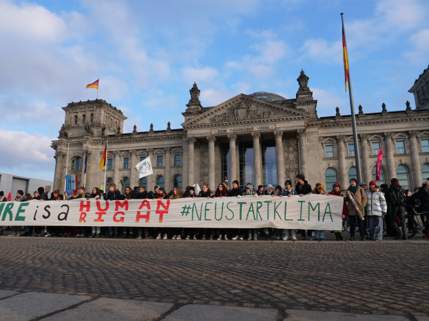 Gruppe von Menschen vor dem Reichstagsgebäude in Berlin, die eine Fahne mit der Aufschrift "Wir sind ein Menschenrecht" halten, mit den architektonischen Details des Gebäudes und umgeben von Flaggen unter einem bewölkten Himmel.