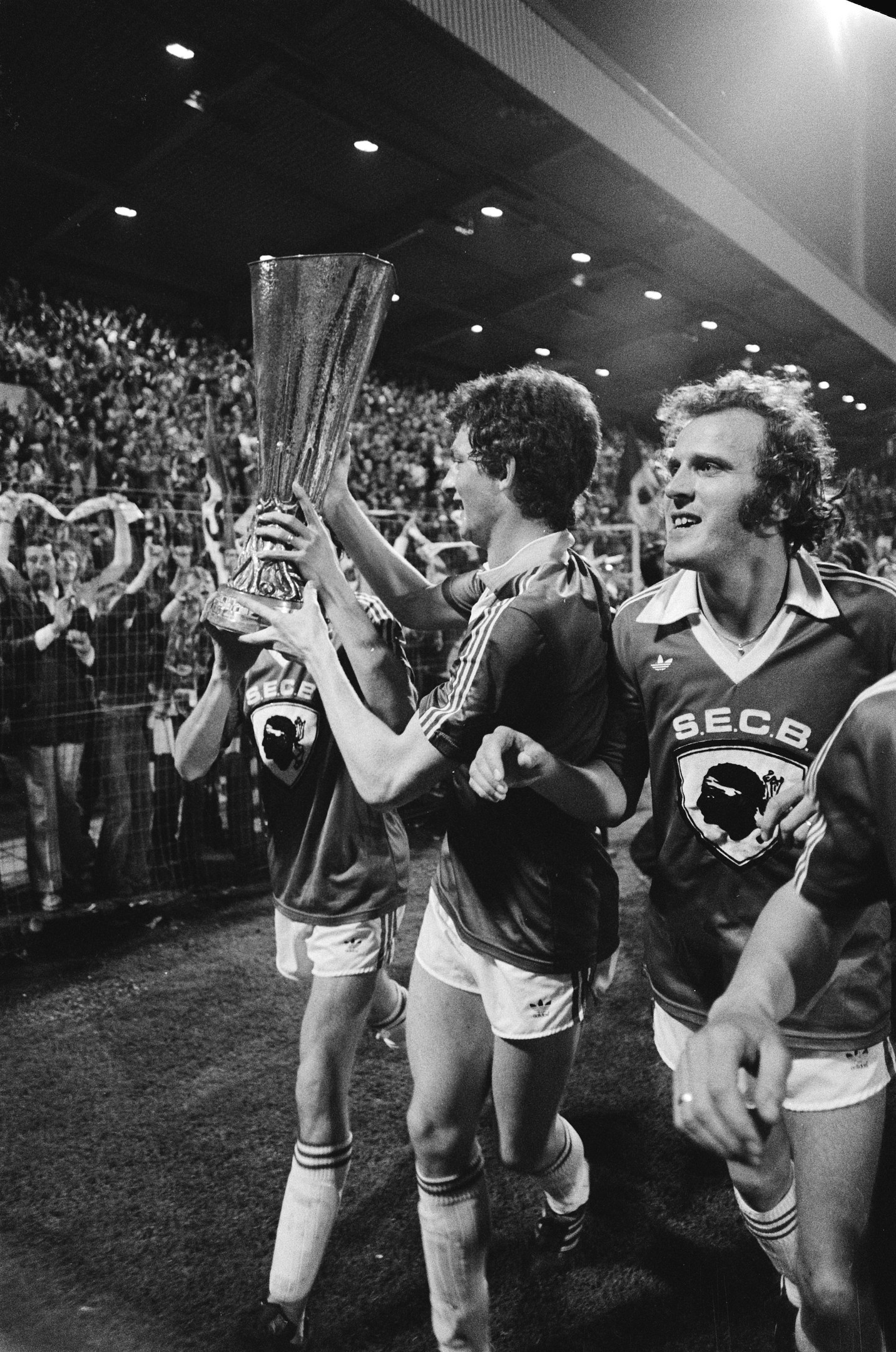 Schwarzes Foto von Fußballspielern, die mit einem Pokal auf einem Feld feiern, umgeben von einer jubelnden Menge hinter einem Zaun unter Stadionbeleuchtung.