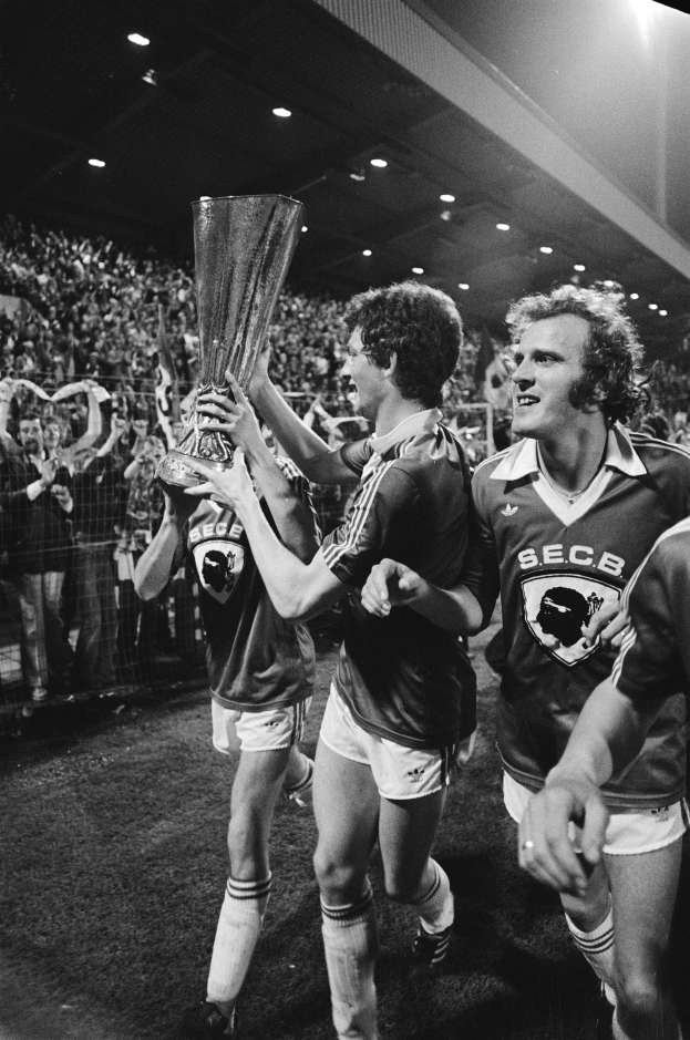 Schwarzes Foto von Fußballspielern, die mit einem Pokal auf einem Feld feiern, umgeben von einer jubelnden Menge hinter einem Zaun unter Stadionbeleuchtung.