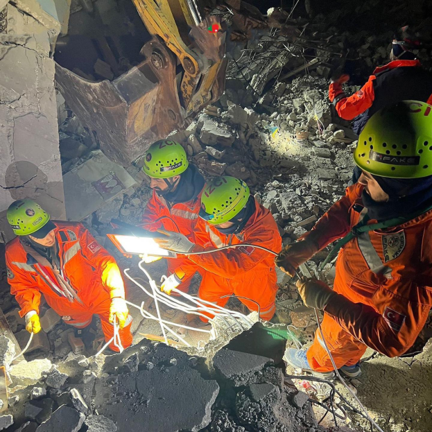 Eine Gruppe von Menschen in orangefarbenen Uniformen und grünen Helmen, die auf Schutt arbeiten, mit einem Kran im Hintergrund, der Rettungsgeräte und Materialien hölt.
