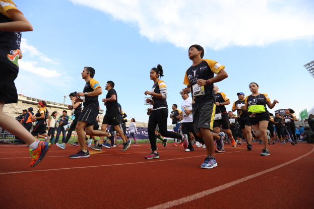 Gruppe von Menschen, die auf einer Laufbahn laufen, mit Zuschauern im Hintergrund, vor Gebäuden und einem klaren blauen Himmel.