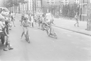 Schwarzes und weißes Bild von Kindern, die Scooter die Straße entlangfahren, mit einem geparkten Auto auf der linken Seite, Gebäuden, Bäumen, Pfählen und einem Zaun im Hintergrund.