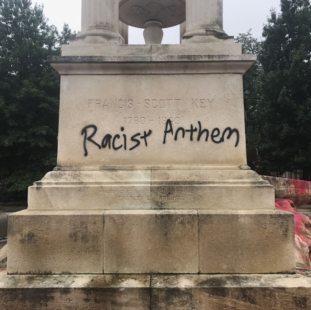 Ein Denkmal mit der Inschrift 'Rassistischer Anthem' vor einem Hintergrund aus Bäumen und einem klaren Himmel.