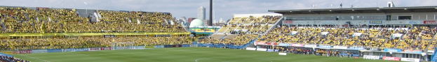 Ein großes Stadion mit Zuschauern, die ein Fußballspiel auf saftigem Grün betrachten, umgeben von Werbetafeln, mit Gebäuden, Pfählen und einem klaren blauen Himmel im Hintergrund.