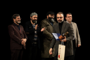 Eine Gruppe Männe steht auf einer Bühne, einer hält eine Tasche und einen Pokal, mit einem dunklen Hintergrund, der die Eröffnung der Berliner Filmfestspiele suggeriert.