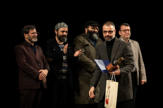 Eine Gruppe Männe steht auf einer Bühne, einer hält eine Tasche und einen Pokal, mit einem dunklen Hintergrund, der die Eröffnung der Berliner Filmfestspiele suggeriert.